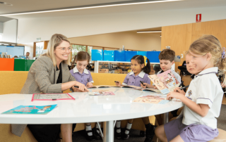 A photo of a teacher speaking to primary school students with books smiling in a very modern classroom facility