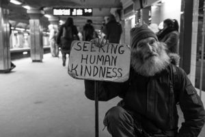 A homeless man holds a sign saying 'Seeking Human Kindness'